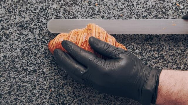Baker in black gloves cuts fresh golden croissant in half on stone countertop. Bakery. Top view. High quality 4k footage
