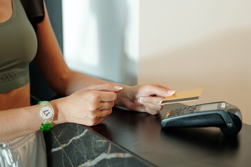 Hands of young woman holding credit card over payment terminal on reception counter in hotel or leisure center lounge