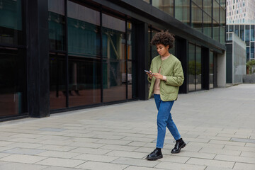 Horizontal shot of curly haired woman enjoys free time in city holds mobile phone chats online walks in urban setting wears jacket jeans and shoes strolls at urban setting checks notification