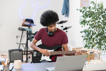 Diligent guy playing electric guitar in front of laptop during online lesson or masterclass in home environment