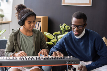 Cute biracial girl in headphones repeating after her music teacher touching keys of piano keyboard - Powered by Adobe