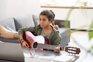 Serious schoolgirl with guitar sitting by table in front of laptop and listening to advice of...