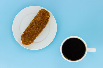 Eclair decorated with sweet crumb in a white plate and cup of black coffee isolated on a pale blue background. Still life with french pastries and coffee. Top view