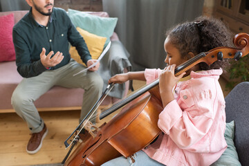 Cute little biracial girl gliding fiddlestick across cello strings while sitting in front of her music teacher © pressmaster