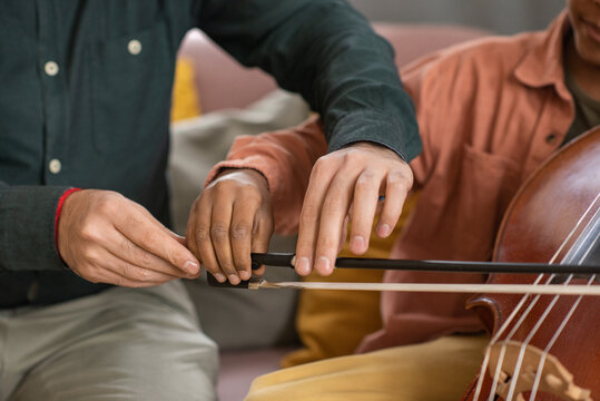 Hands of music teacher and his pupil playing cello while young man holding fiddlestick and helping little boy to glide it