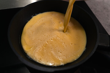 Whipped chicken eggs for omelet are poured from a metal bowl into a black skillet on an induction...