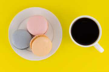 Still life with cup of black coffee with assorted colorful macarons on plate isolated on yellow background. Almond French macaroon cookies and espresso cup. Top view