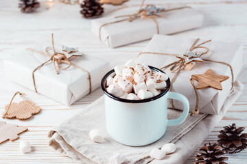 Cocoa and marshmallows in a mug next to craft gifts on a white wooden table. Hot sweet drinks