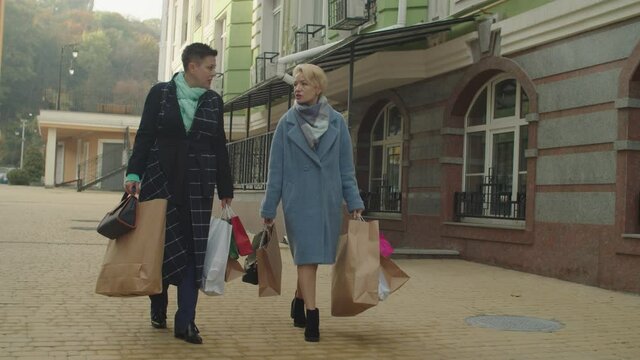Stylish female friends walking down city street, carrying many paper shopping bags outdoors. Middle-aged women shopping, enjoying pastime at retirement