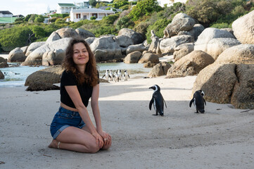 Happy young woman sits on Boulders beach with penguins