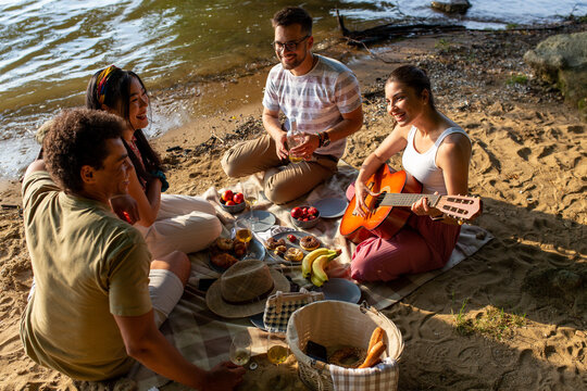 A group of diverse friends playing guitar enjoys music and food on the beach. Picnic time.