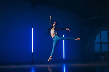 Young woman in casual style - jeans and leather jacket doing ballet in modern studio. Attractive ballerina practices in choreography alone