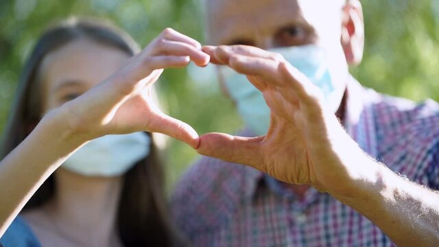 Old man and teenage granddaughter in masks connect hands in heart symbol indicating unity after weakening covid restrictions in garden