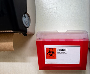 Red biohazard sharps waste container mounted to the wall of a public restroom - obrazy, fototapety, plakaty