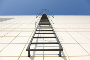 Long metal staircase on the gray modern facade of an industrial building, warehouse or shopping center against the blue sky. Fire fighting equipment concept