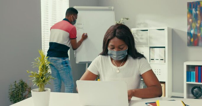A busy man stands at the blackboard holding a tablet, looking for the right graphic, preparing a diagram, a beautiful woman sits at her desk entering data into the system needed to create the diagram