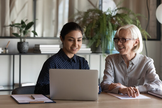 Old age female employee share idea discuss project with young indian colleague at corporate office. Millennial mixed race woman mentor explain task on computer teach work in app mature lady intern
