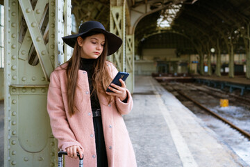Fototapeta na wymiar Beautiful young woman waiting for train at train station for travel in autumn. With phone in hand, he communicates via video communication or takes selfie on smartphone. In pink coat. Travel concept.