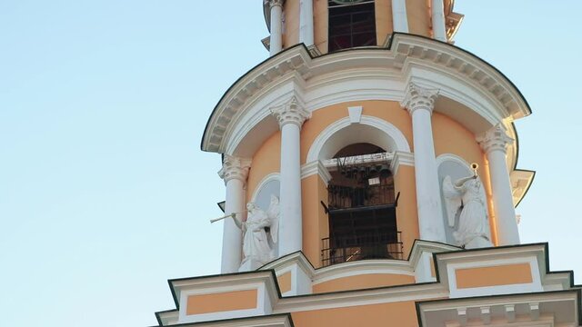 bell tower of peach color with white edging, the sky is blue, evening, sunset. the bell ringer rings the bells, white anegles on the wall