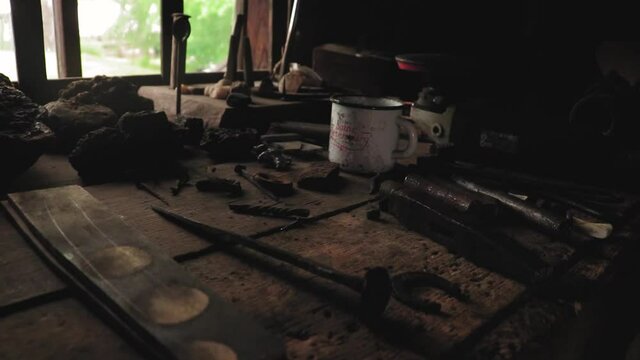 blacksmith work table, white iron mug, blacksmith tools on a wooden table, metal parts for blacksmiths, natural light from behind the window