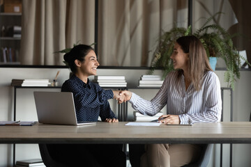 Two smiling diverse young women corporate workers shake hands satisfied with successful cooperation making good deal. Millennial indian female employee handshake colleague appreciate for help in work