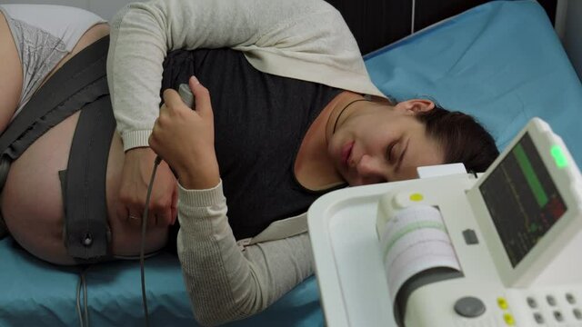 Young Pregnant Woman On Hospital Couch During Medical Control Cardiotocography. Visit Gynecologist Doctor At Clinic For Pregnancy Consultant. Examine Expectant Belly Baby Mother Healthcare Check Up