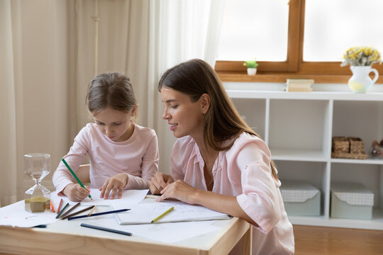 Happy caring beautiful young mother or nanny drawing teaching little smiling kid girl drawing in paper album, involved in creative early development activity together at home, daycare concept.