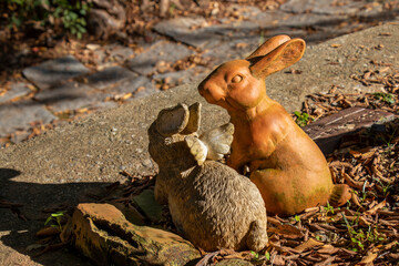 Two weathered terracotta rabbit figurines, one with angel wings, sit in slanting sunlight at the corner of a rustic raised bed along a cobblestone garden pathway scattered with leaves.
