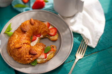 Close-up. Delicious ruddy croissant smeared with butter, fresh strawberries and a cup of coffee on a turquoise background. French breakfast. Recipe book, cooking blog, café, restaurant.