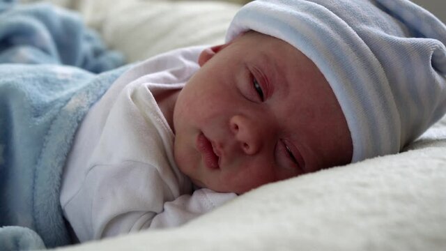 Close-up Newborn Baby Face Portrait Early Days Sleeping Sweetly On Tummy Blue White Background. Child At Start Minutes Of Life on Hat. Infants, Childbirth, First Moments Of Borning, Beginning Concept