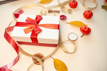 Autumn composition in the form of a gift box with a red ribbon, small pumpkins, candles and gold ribbons. Beige background.