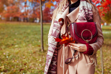 Naklejka premium Close up of stylish woman holding burgundy purse wearing coat in autumn park. Fall female clothes, accessories. Fashion