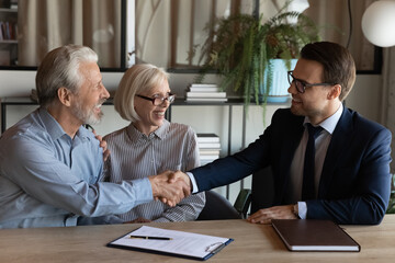 Shaking hands. Smiling young man real estate broker handshaking with older family couple after buying selling property. Spouses retirees thank bank agent for help after signing loan mortgage agreement - obrazy, fototapety, plakaty