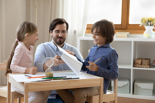 Smiling millennial father teaching small children son daughter writing letters or drawing pictures in paper album, sitting at table with sand clock, enjoying spending creative time together at home.