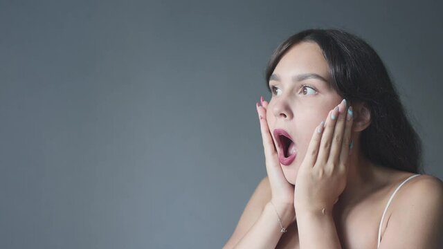 expressive caucasian brunette female 20 saying wow wondering on grey background copy space. portrait young woman chocked surprised mouth opened hands cheeks big eyes wide. emotions advertising concept