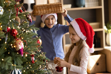 Cheerful small children brother sister having fun decorating evergreen fir tree, playing having fun preparing for Christmas winter holidays, enjoying entertaining festive activity together at home.