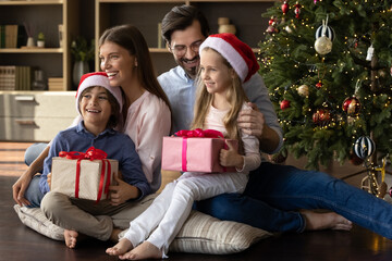 Fototapeta na wymiar Happy bonding young family couple sitting on pillows on floor in living room with laughing little kids on laps near decorated festive Christmas tree, holding wrapped boxes with presents gifts.