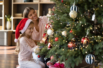Happy pretty small adorable child daughter in red hat sitting on warm heated floor, decorating Christmas fir tree with happy caring loving young mother, preparing for New Year winter holidays at home.