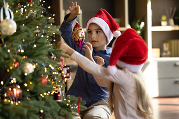 Happy cute little kid boy decorating Christmas tree with beautiful baubles together with small sister, enjoying designing branches with lights and decor, involved in preparing for New Year holidays.