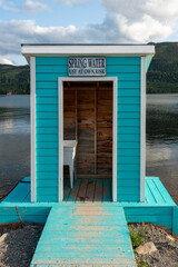 A teal green color wooden watershed with white trim near a pond. The doorway is open and there's a white plastic sink inside. There's a sign over the door with the text spring water use at own risk. 