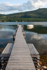 A long wooden boat wharf with a white boat moored. The plank pier juts out into the ocean on all three sides with tree covered land in the background under blue sky with clouds.  