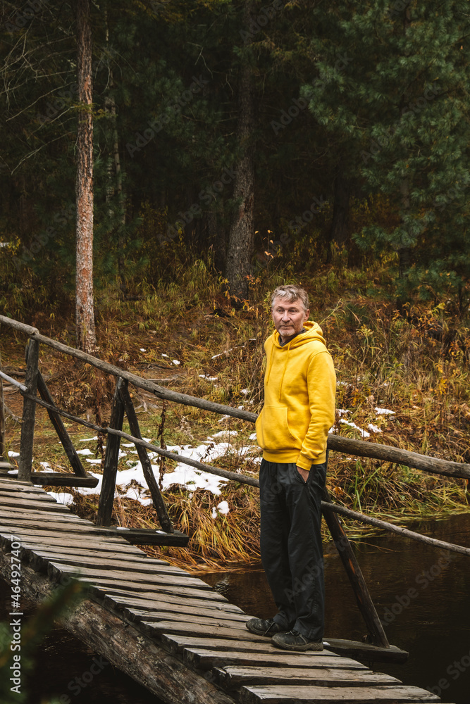 Wall mural A middle-aged traveler in yellow hoodie stands on wooden bridge over a river in the forest, enjoying beautiful autumn day outdoors. Local travel and weekend hikes. Eco tourism. Vertical. - Wall murals