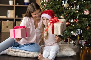 Obraz na płótnie Canvas Happy young mother embracing small laughing kid daughter, having fun holding unwrapped gift boxes, sitting on cozy pillows on floor near decorated Christmas tree, New Year holidays family celebration.