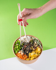 A woman's hand with chopsticks over a salad poke bowl and tuna, carrot, cucumber and pineapple with a green background.