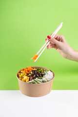 A woman's hand with chopsticks over a salad poke bowl and tuna, carrot, cucumber and pineapple with...
