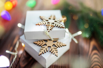 Two white gift boxes decorated with wooden snowflakes. Packing gifts for Christmas and New Year on a beautifully decorated wooden table with colorful garlands and lights.