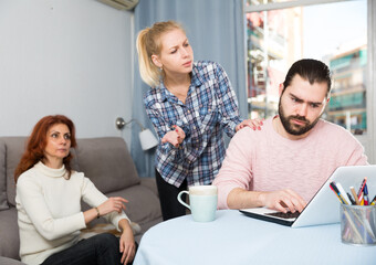 Upset young man working at laptop, young sister and mother on background