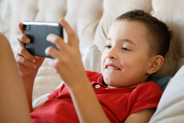 The boy is playing games on his smartphone, lying on the couch at home.