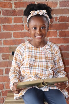 Cute little black girl smiling holding open book vertical brick background