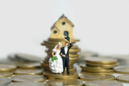 Housing for newlywed concept miniature people, toys photography. Bride and groom standing above coin pile in front of mini house isolated on white background.